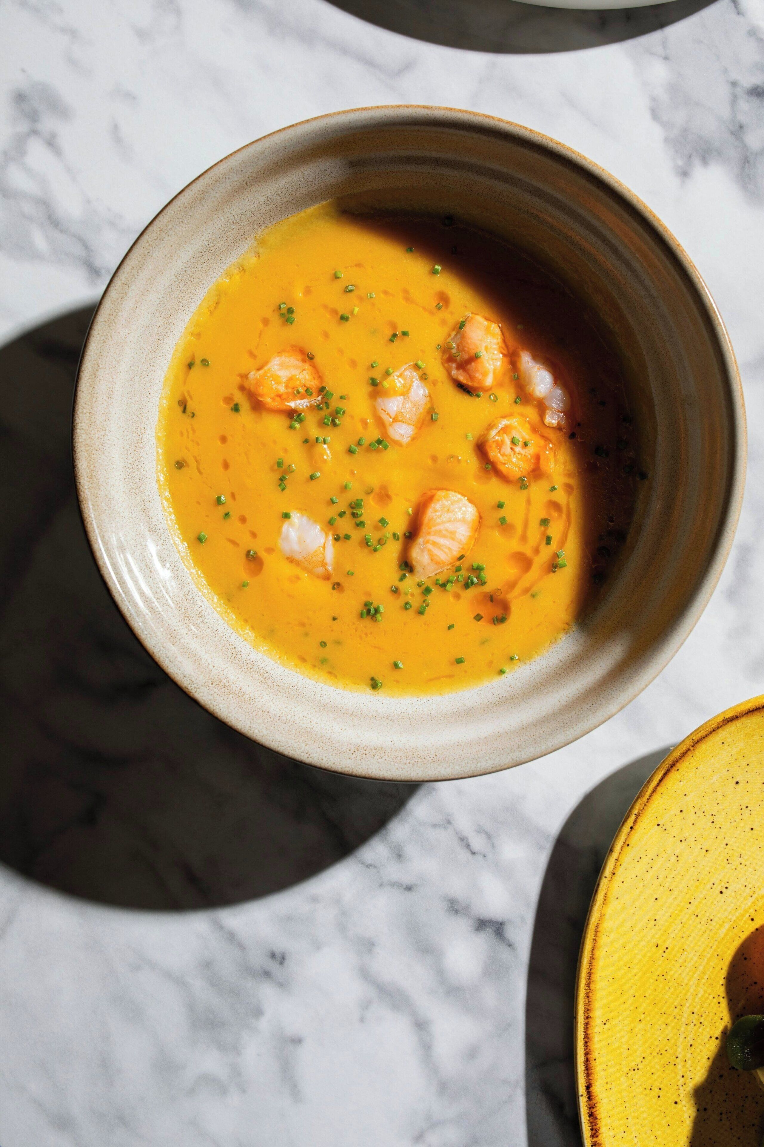 A bowl of soup with shrimp and herbs on a marble surface.
