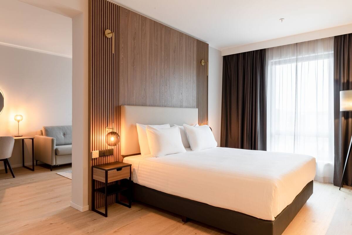 Una habitación de hotel moderna con una cama grande, un panel de madera decorativo, una mesita de noche con lámpara, un sofá y vistas de la ciudad a través de amplios ventanales.
