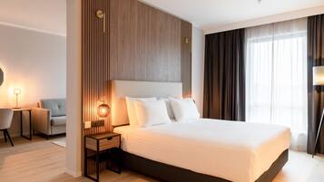A modern hotel room with a large bed, a wooden accent wall, a bedside table with a lamp, a sofa, and a view of the city through large windows.