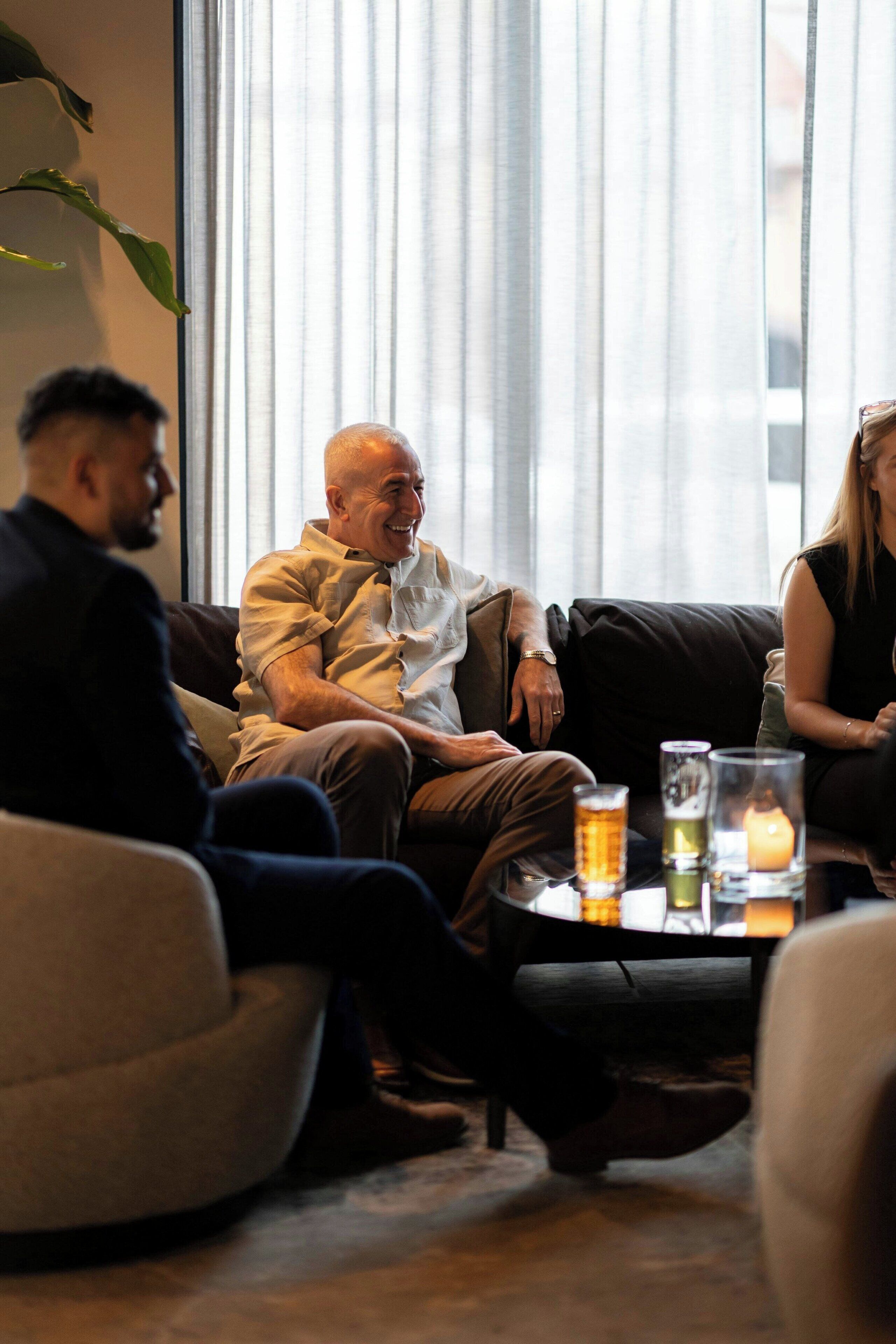 Eine Gruppe von Personen sitzt auf einer Couch in einem Raum mit einem großen Fenster, einem Tisch mit Getränken und einer Kerze.