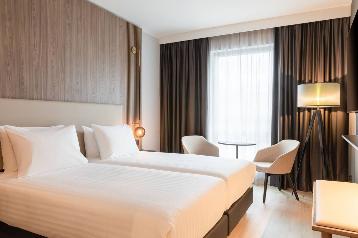Habitación de hotel con dos camas, un escritorio, una silla, una lámpara y vistas al exterior a través de amplias ventanas.