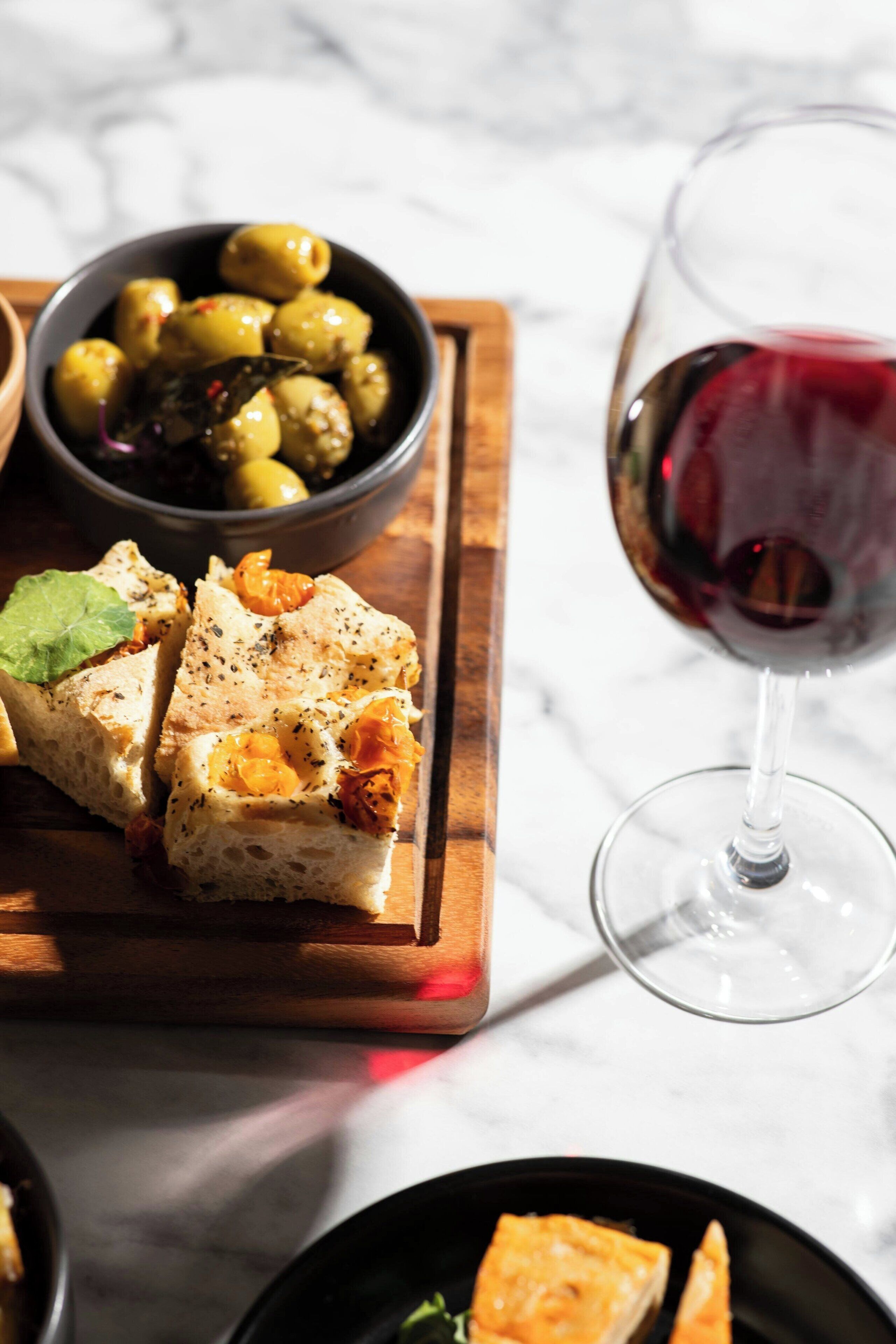 Une planche en bois sur laquelle on a disposé une tranche de pain garnie d’herbes et de fromage, un bol d’olives et un verre de vin rouge.