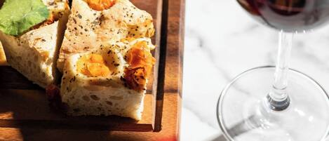 A wooden board with a slice of bread topped with herbs and cheese, a bowl of olives, and a glass of red wine.