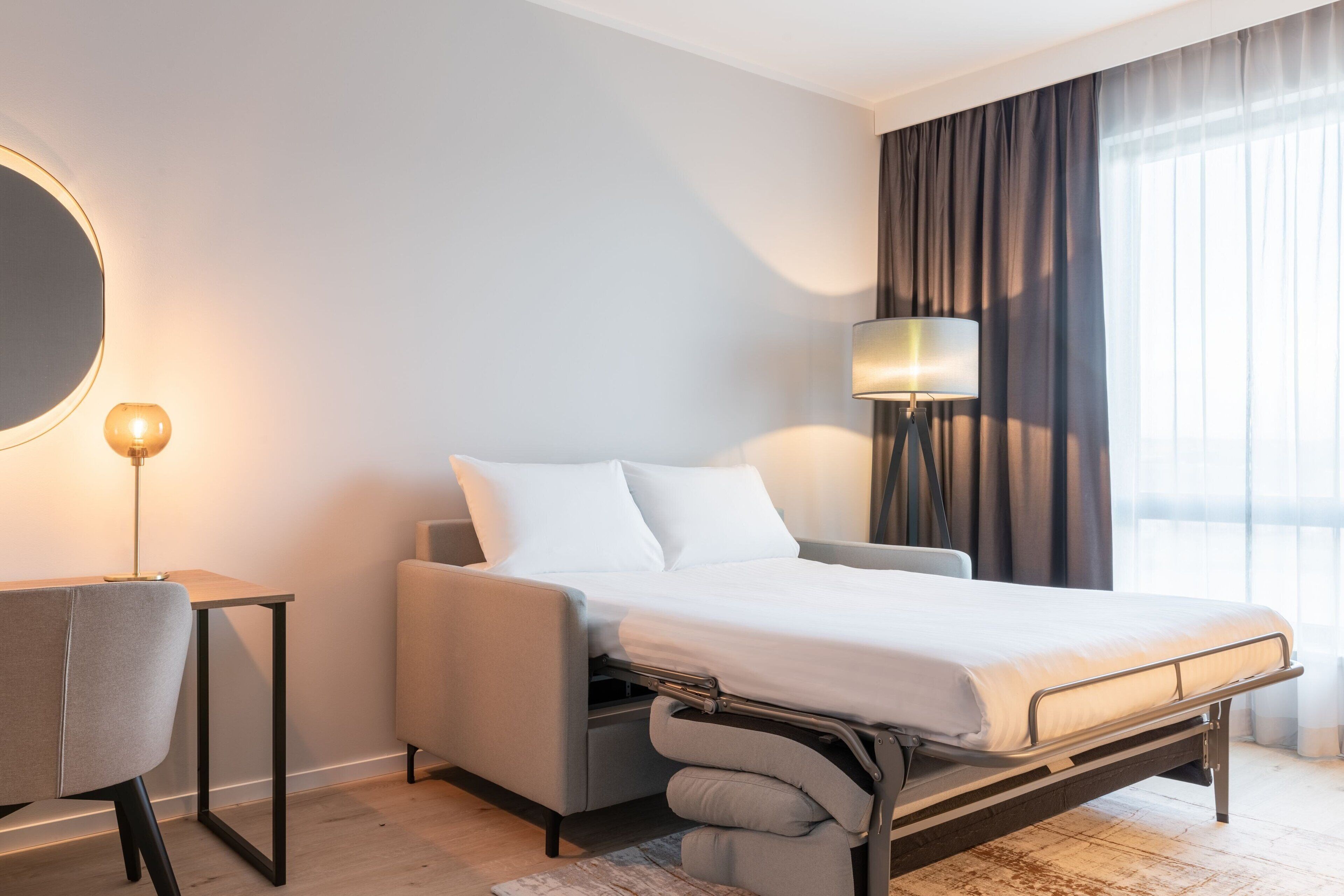 A modern hotel room with a bed, a desk, a chair, a lamp, and a large window with curtains.