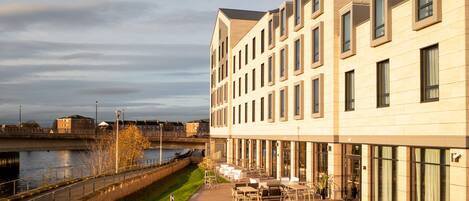 A modern building with a riverside patio area, featuring outdoor seating and a view of the water.
