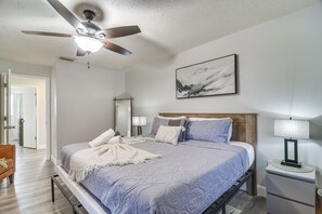 Quarto com cama, ventilador de teto, mesa de cabeceira com abajur, quadro pendurado na parede e porta que dá para outro cômodo.