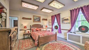 Un dormitorio con cama de madera, colcha estampada en rojo y beige, mesita de noche con lámpara, cómoda, ventana con cortinas moradas y radiador fijado a la pared.