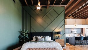 A modern bedroom with a tufted headboard, a wooden bench, a green accent wall, and a kitchen area with bar stools. - Tailor Loft Studio| Living in the Heart of Knox (Knoxville)