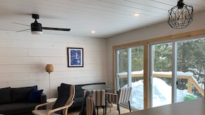 A modern living room with a sofa, chairs, a glass table, and a view of a snowy deck through large windows. - Perfect 4-season cottage for year-round joy (Whitestone)