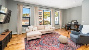 Una sala moderna con un sofá seccional color beige, un tapete estampado, un sillón gris y un televisor de pantalla plana montado en la pared.