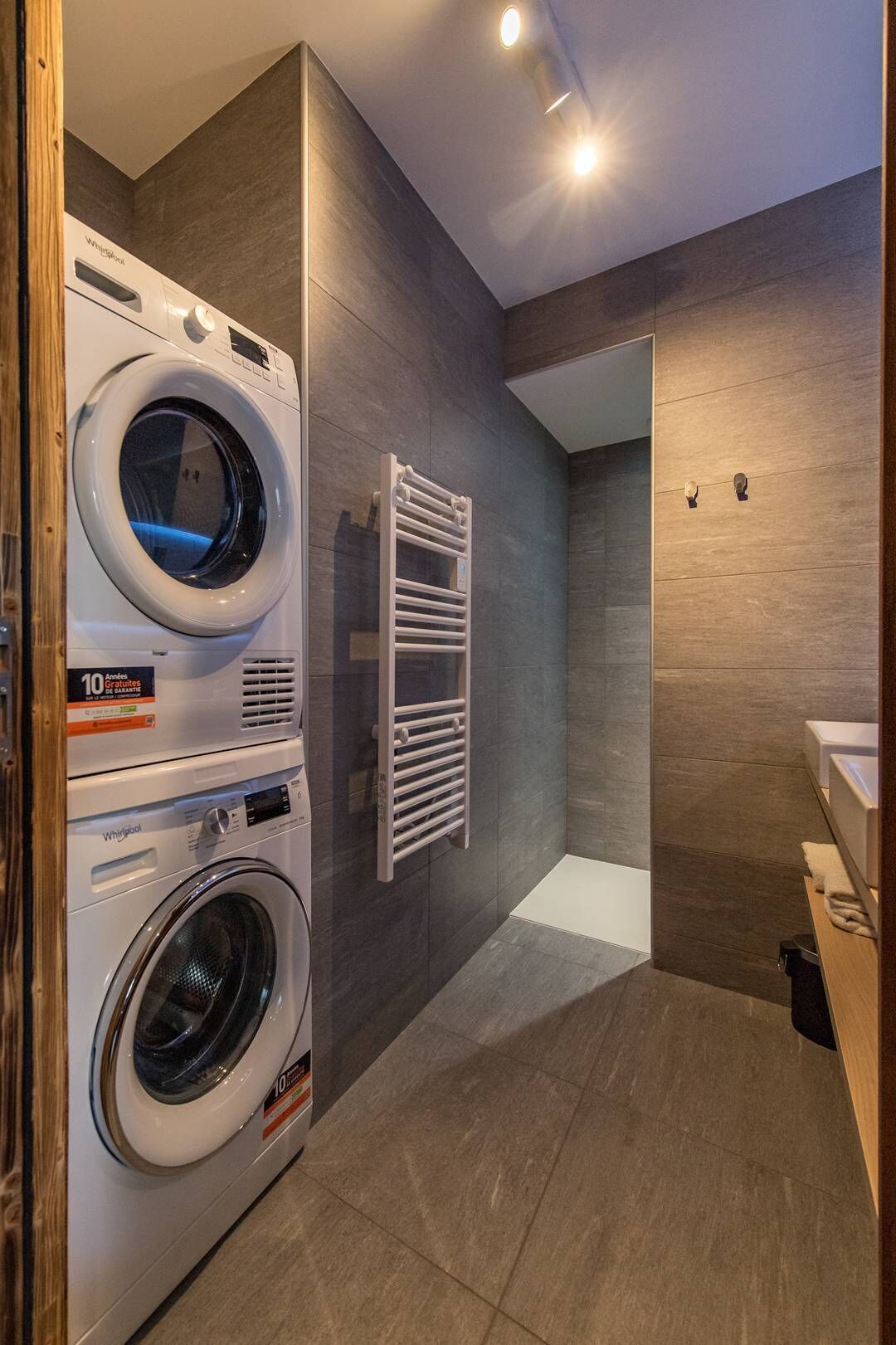 A modern laundry room with two stacked front-loading washing machines, a built-in towel rack, and a wooden floor.