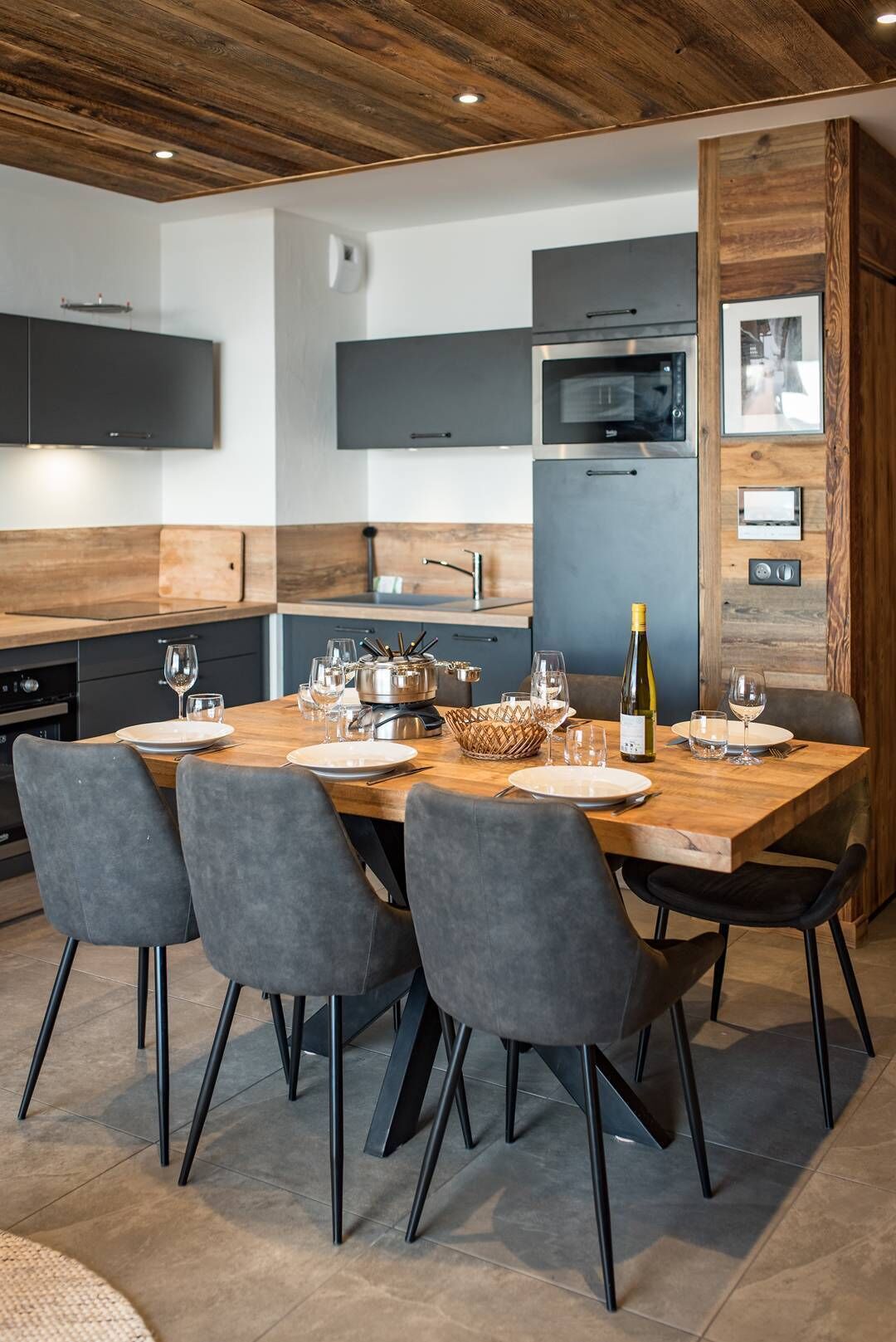 Une cuisine moderne avec une table à manger en bois, préparée pour accueillir six personnes, et équipée d’un four à micro-ondes, d’un four traditionnel et d’une bouteille de vin.