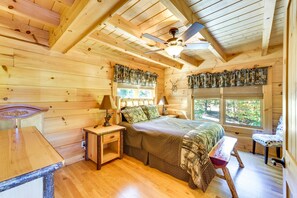 Een houten slaapkamer met een bed, nachtkastjes, een bankje, een plafondventilator en een raam met uitzicht op bomen.