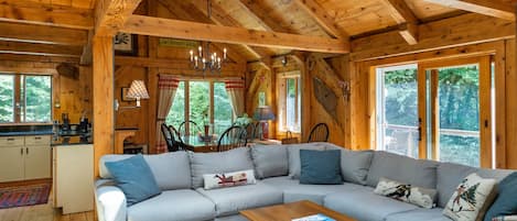 A log cabin interior with a large gray sectional sofa, a wooden dining table, and a kitchen area with a window overlooking greenery.