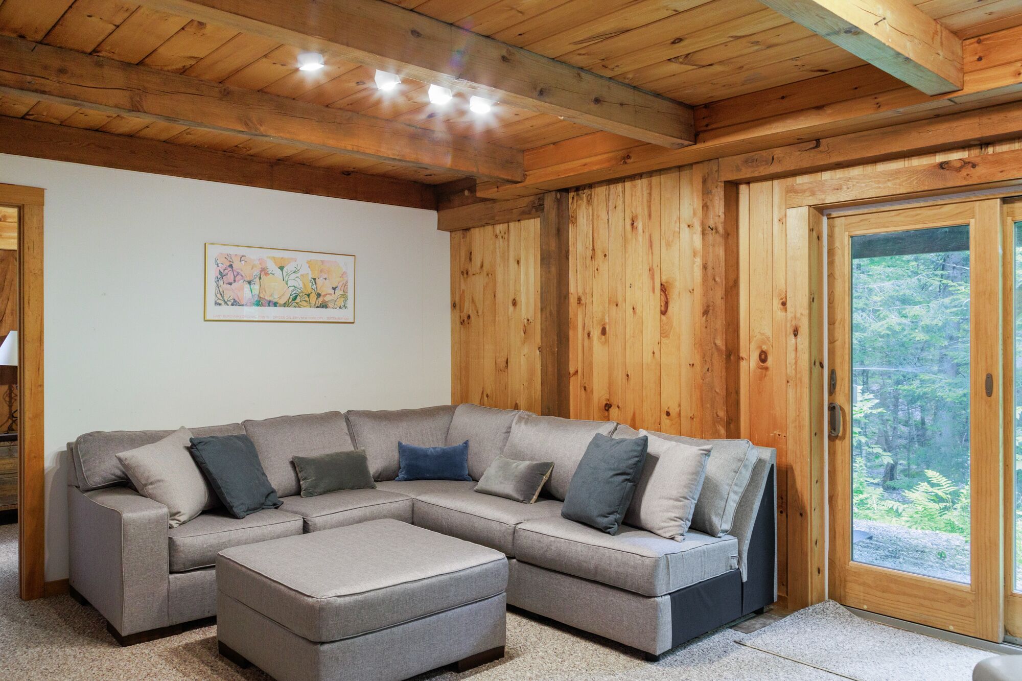 Un salon chaleureux avec un canapé d’angle et un pouf, des murs en bois, une grande fenêtre donnant sur la forêt et une œuvre d’art encadrée accrochée au mur.
