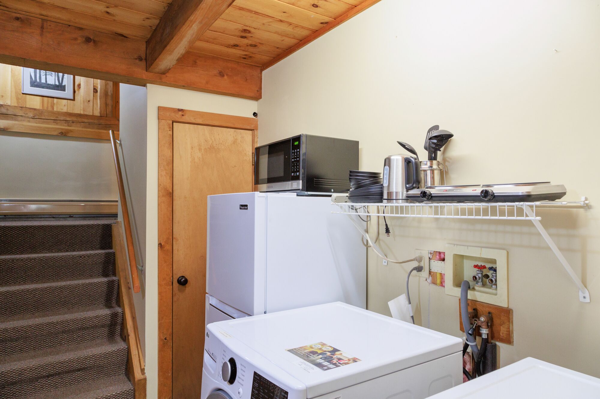 Cocina compacta con microondas, cafetera y un refrigerador pequeño. Se observa una puerta de madera y una escalera que sube a otra habitación.