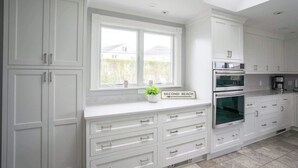 Une cuisine moderne avec des meubles blancs, un four encastré et une fenêtre donnant sur une maison.