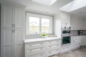 A modern kitchen with white cabinetry, a built-in oven, and a window with a view of a house. - Beach Haven by STAY Newport (Middletown)