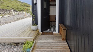 Une terrasse en bois mène à un bâtiment sombre également en bois, doté d’une porte, d’une plante en pot et d’un banc.