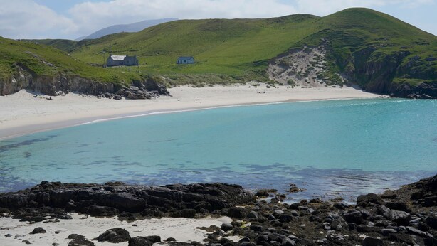 Un paysage côtier avec une plage de sable, des eaux turquoise et limpides, et des collines verdoyantes en arrière-plan.