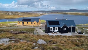Une propriété située sur la côte, comprenant deux cabanes, l’une avec un toit noir et l’autre avec un toit marron, toutes deux équipées de panneaux solaires, et offrant une vue sur une étendue d’eau et des collines.