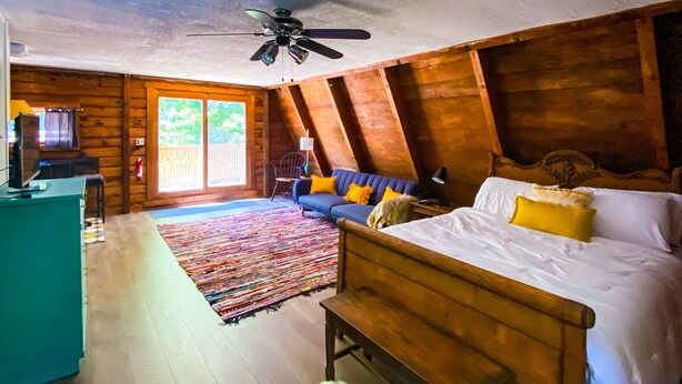 Une chambre avec un plafond en bois, un lit avec des oreillers, un canapé avec des coussins, un banc en bois et un téléviseur posé sur un meuble.