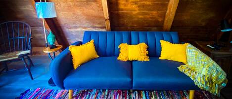 Ein blaues Sofa mit gelben Kissen, ein farbenfroher Teppich und eine Buddha-Statue auf einem kleinen Tisch.