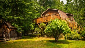 Une cabane en bois dotée d’un balcon, entourée d’une végétation luxuriante et d’un petit abri de jardin.