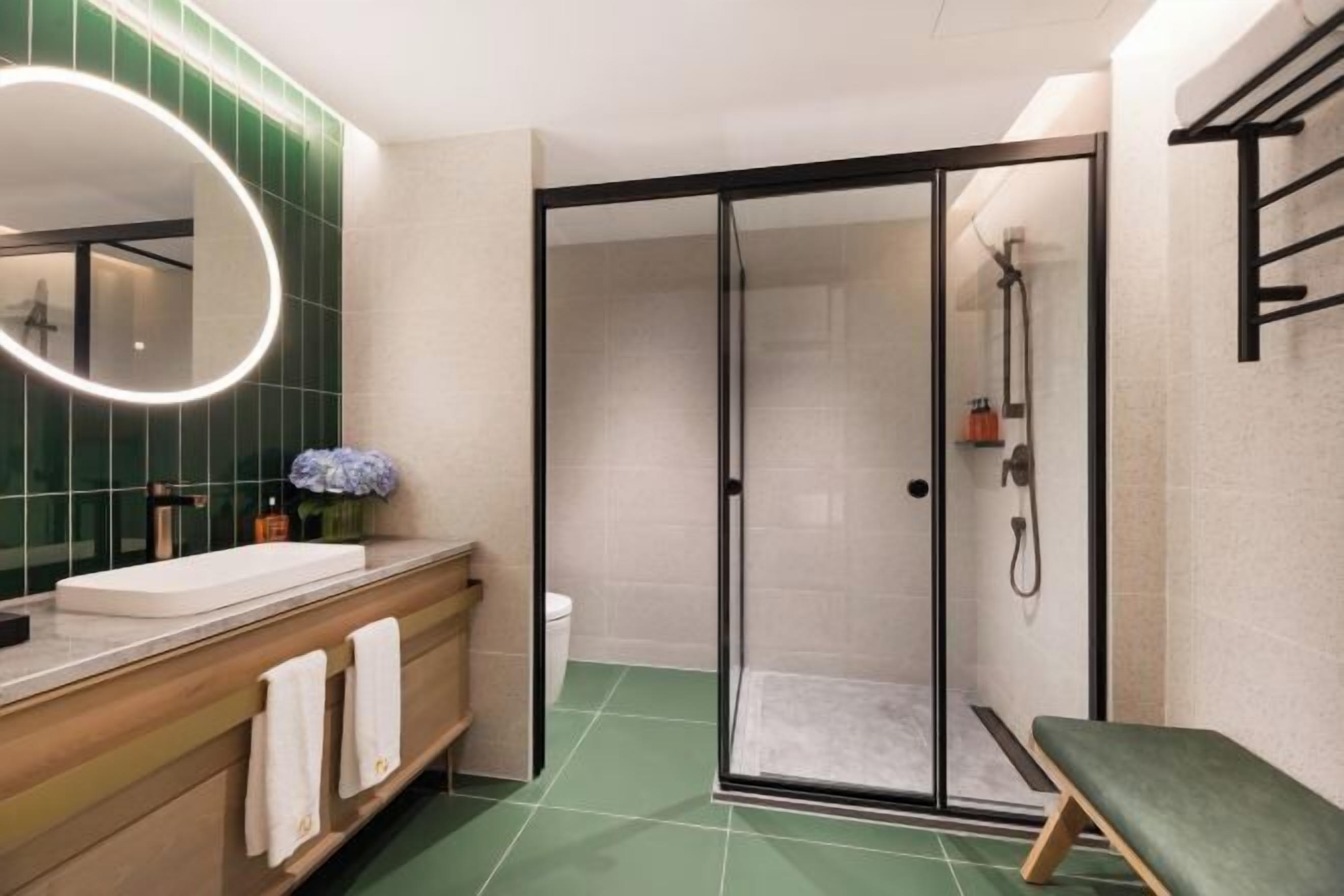 Um banheiro moderno com chuveiro em gabinete de vidro, toucador de madeira com pia branca e piso revestido de azulejos verdes.