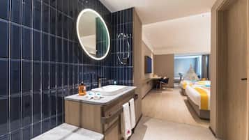 Um banheiro de hotel moderno com um espelho grande, pia e bancada. No fundo, vê-se uma cama com colcha amarela e branca.