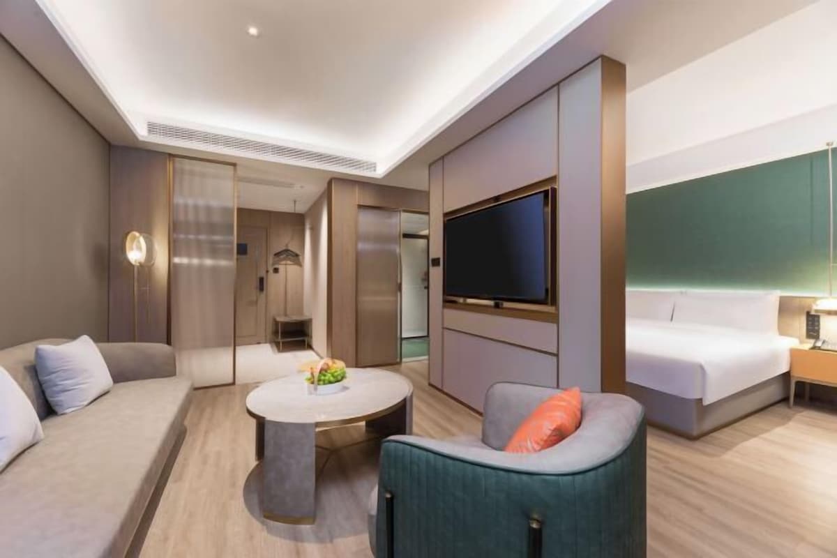 A modern hotel room with a sofa, a round coffee table, a TV mounted on the wall, and a bed with a nightstand.