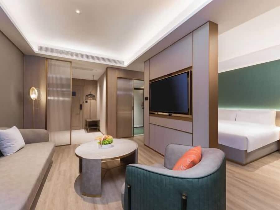 Quarto de hotel moderno com sofá, mesa de centro redonda, televisão fixada na parede e cama com mesa de cabeceira.