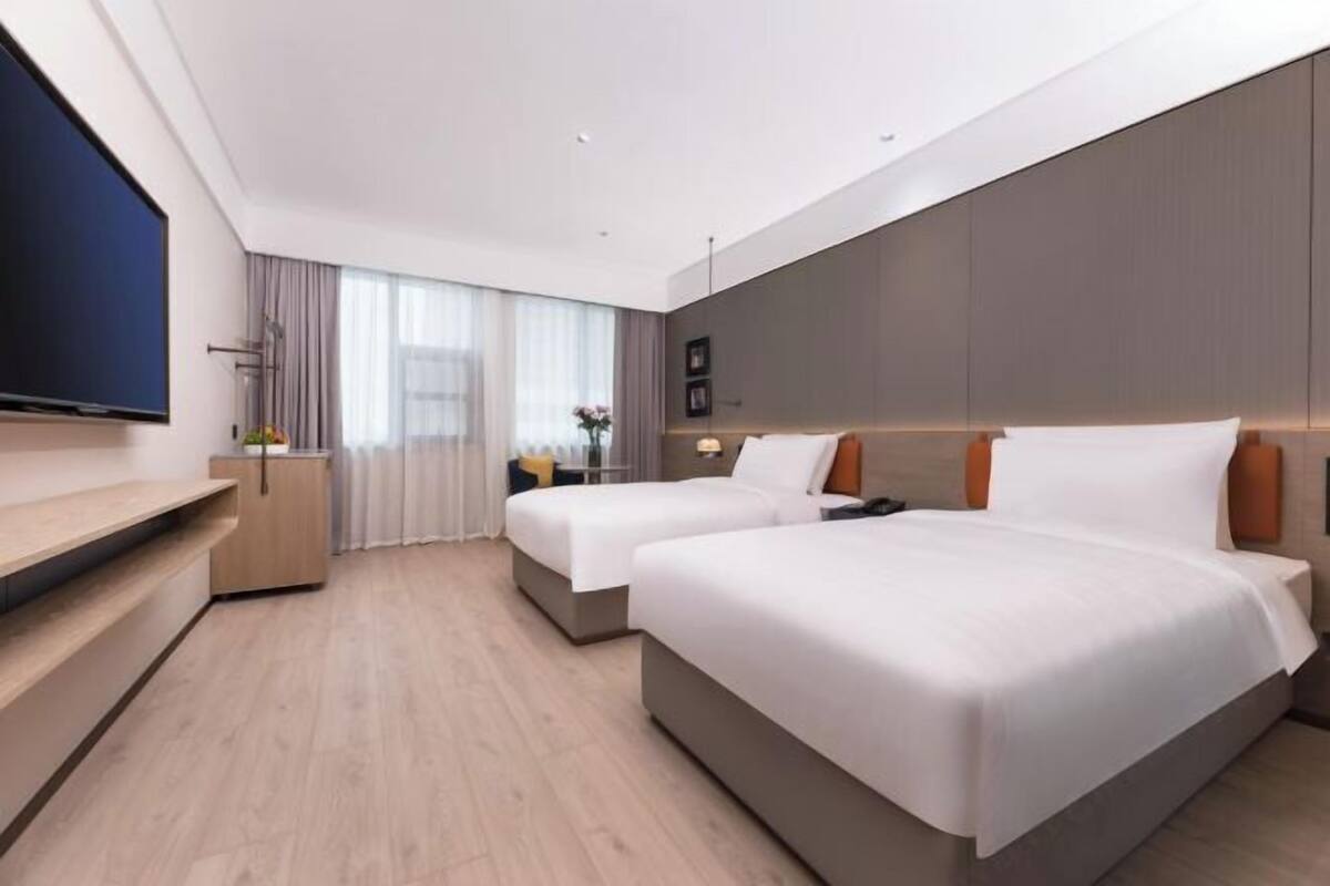 A modern hotel room with two beds, a flat-screen TV, a desk, and a large window with curtains.