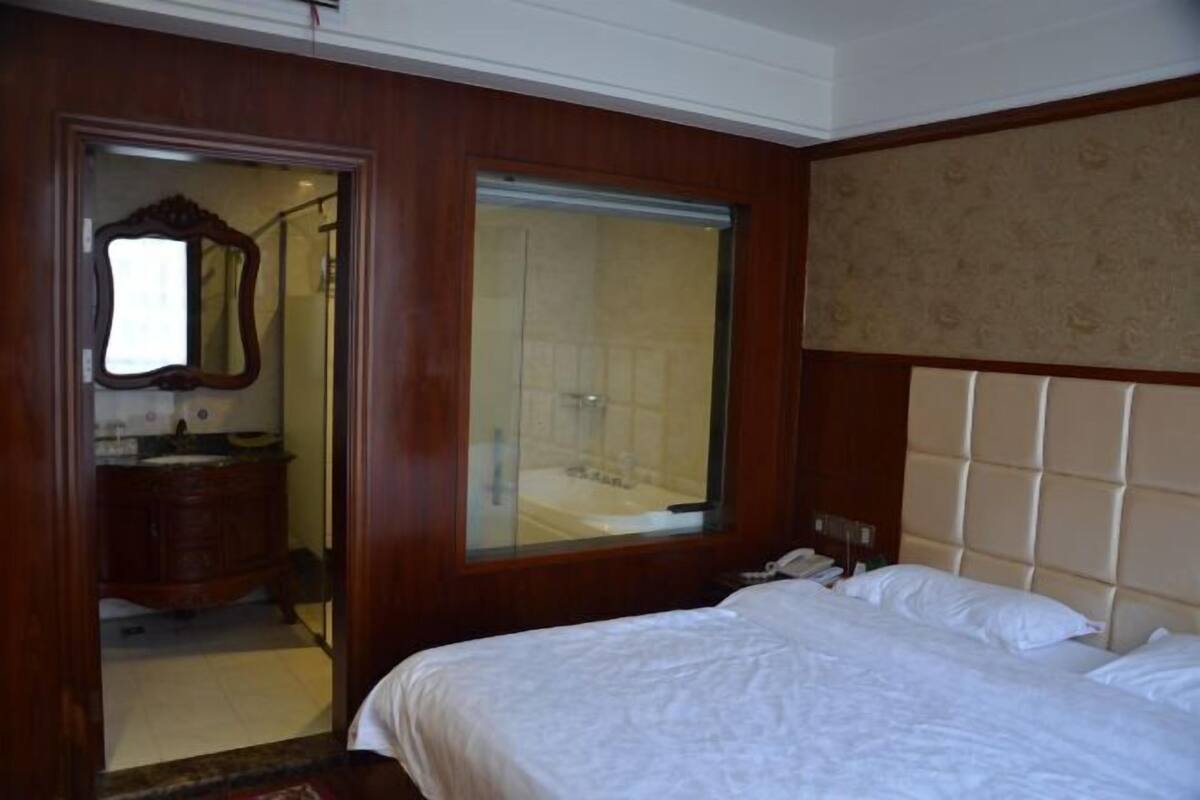 A hotel room with a wooden paneled wall, a large window, a bed with white linens, and a bathroom visible through an open door.