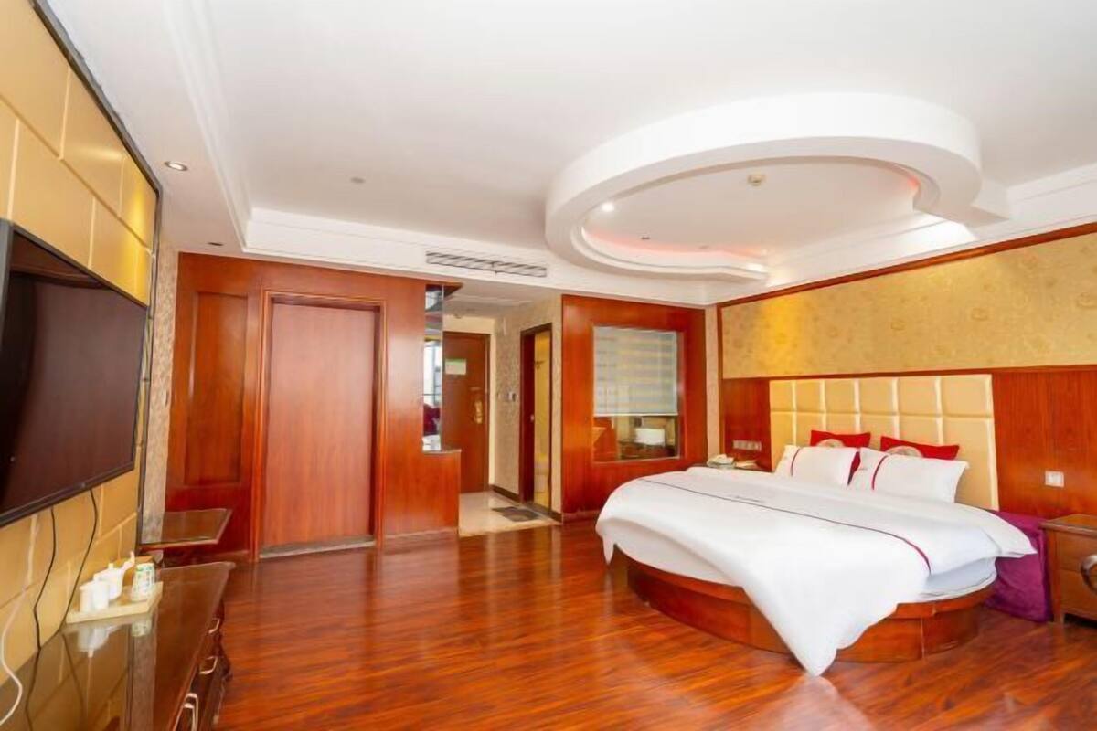 A hotel room with a large bed, a TV, a wooden floor, and a modern ceiling design.