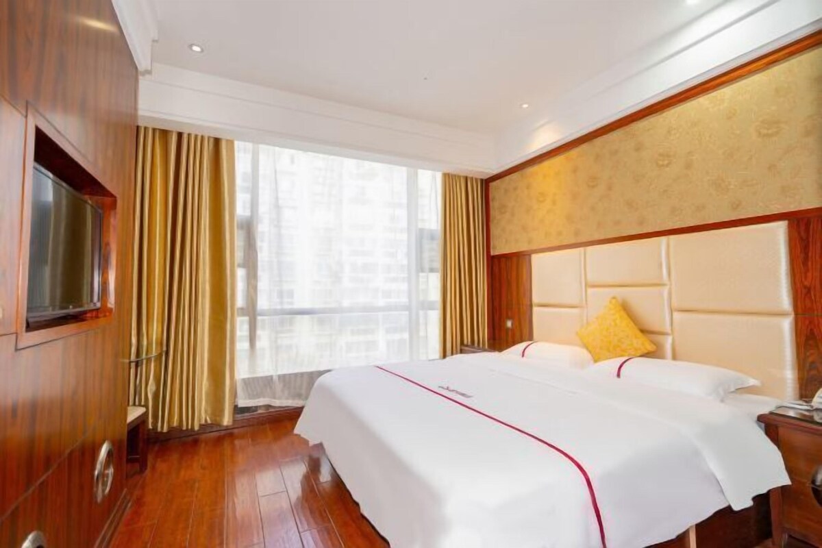 A hotel room with a large bed, a television mounted on the wall, and a window with curtains.