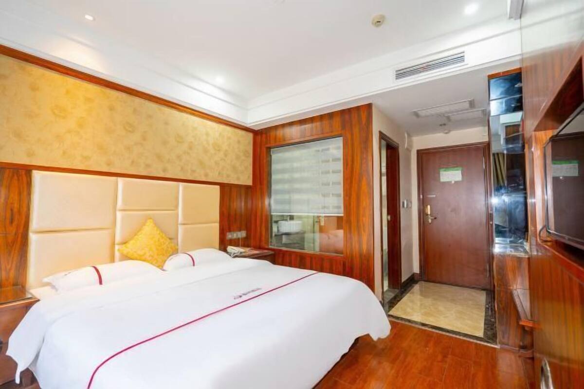 A hotel room with a large bed, wooden headboard, and a glass partition.