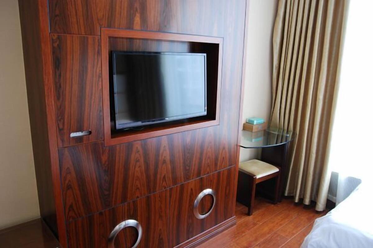 A hotel room with a wooden cabinet, a flat-screen TV, a glass table, a chair, and a bed.