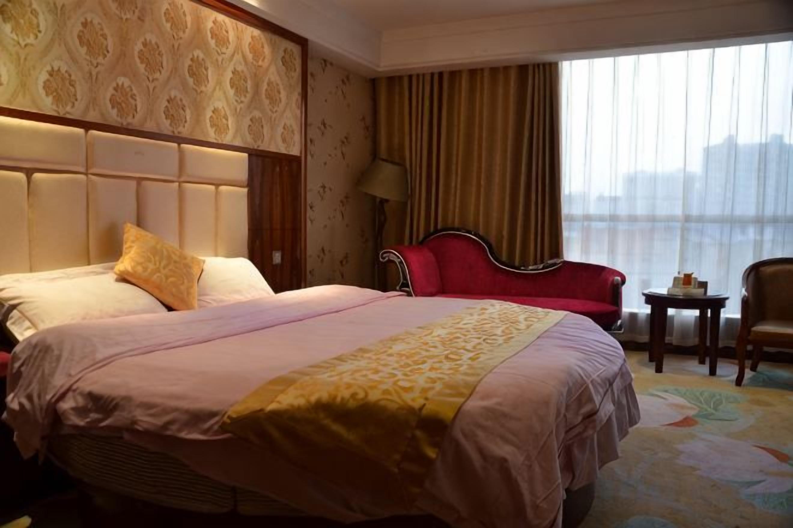A hotel room with a large bed, a red armchair, a small table, and a view of the city through the window.