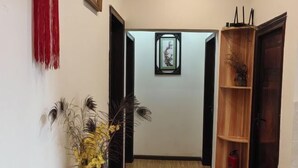 A hallway with a wooden floor, a red decorative hanging, a framed artwork on the wall, and a wooden shelf with a vase and a fire extinguisher.