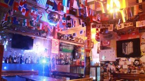 A bar with wooden benches, a variety of bottles on shelves, and a wall decorated with flags and posters.