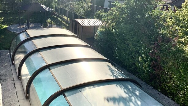 Una piscina amb una estructura de vidre corbada, envoltada de vegetació i edificis residencials al fons.