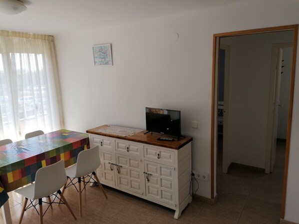 Una habitación con una mesa de comedor, sillas, un televisor sobre un mueble, un cuadro pequeño en la pared y una puerta que da a otra habitación.