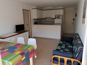 Un espace de vie compact comprenant un coin cuisine, un coin repas avec une nappe colorée et un canapé recouvert d’un tissu à motifs.