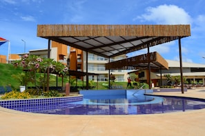 Una piscina con azulejos azules, rodeada por una zona techada con listones de madera, y un edificio con balcones al fondo.