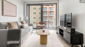Een moderne woonkamer met een grijze bank, een houten salontafel, een zwart tv-meubel en een balkon met uitzicht op een bakstenen gebouw.