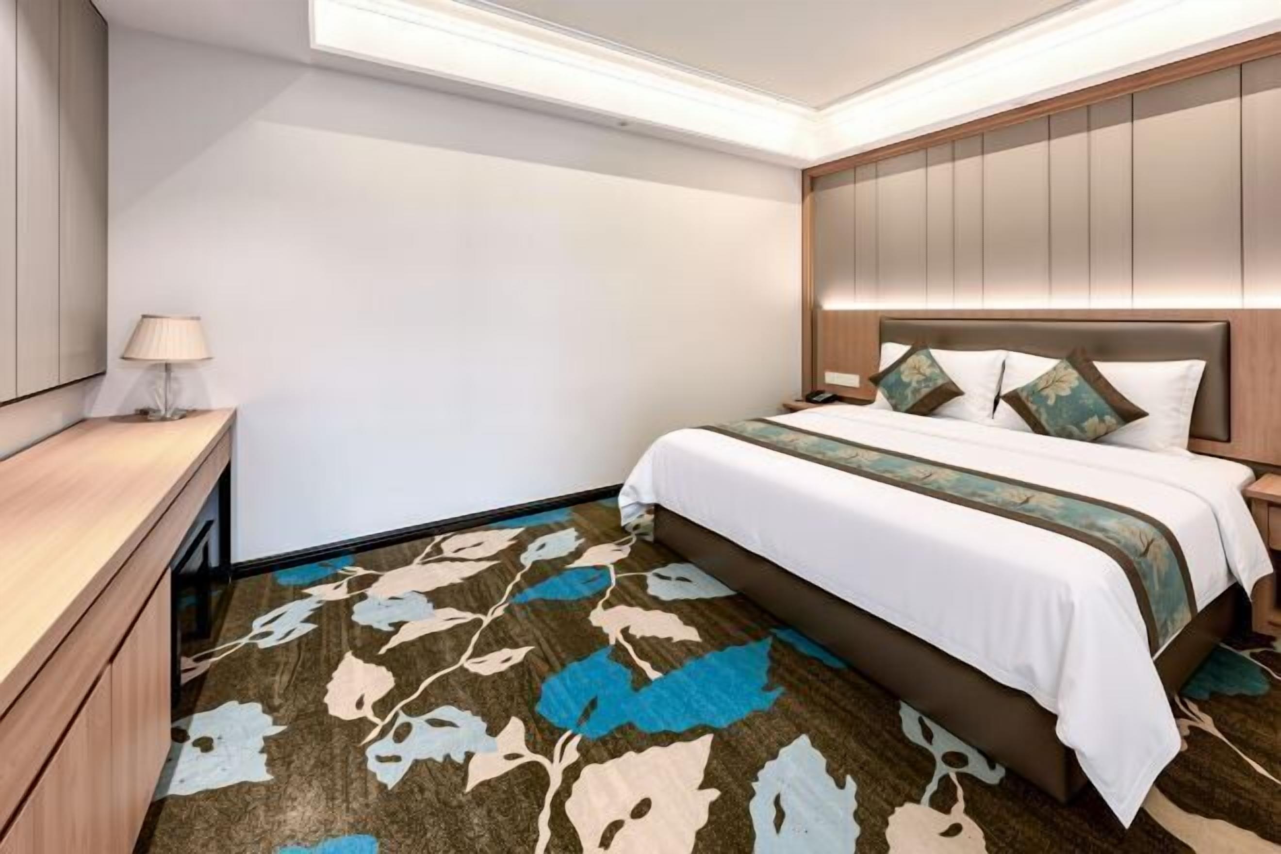 A hotel room with a bed, bedside table, lamp, and a patterned carpet.