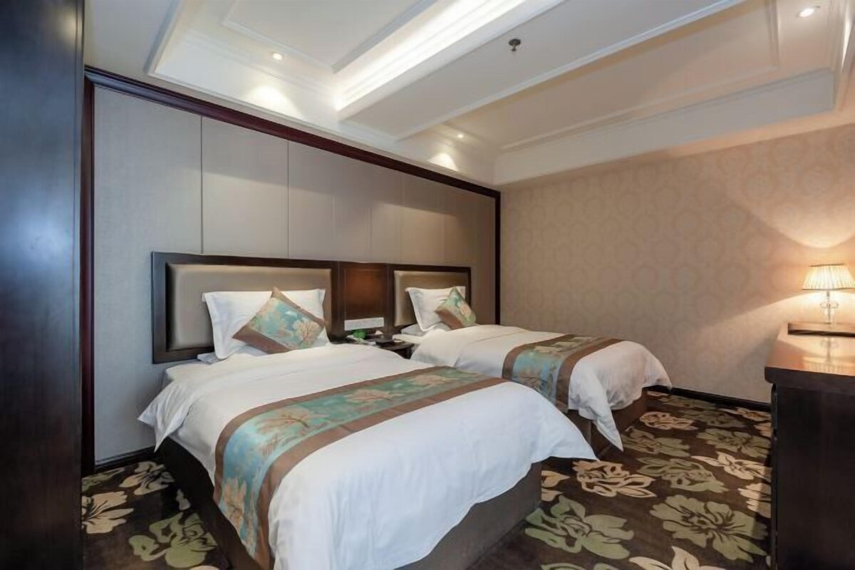 A hotel room with two beds, a wooden headboard, a floral carpet, and a nightstand with a lamp.