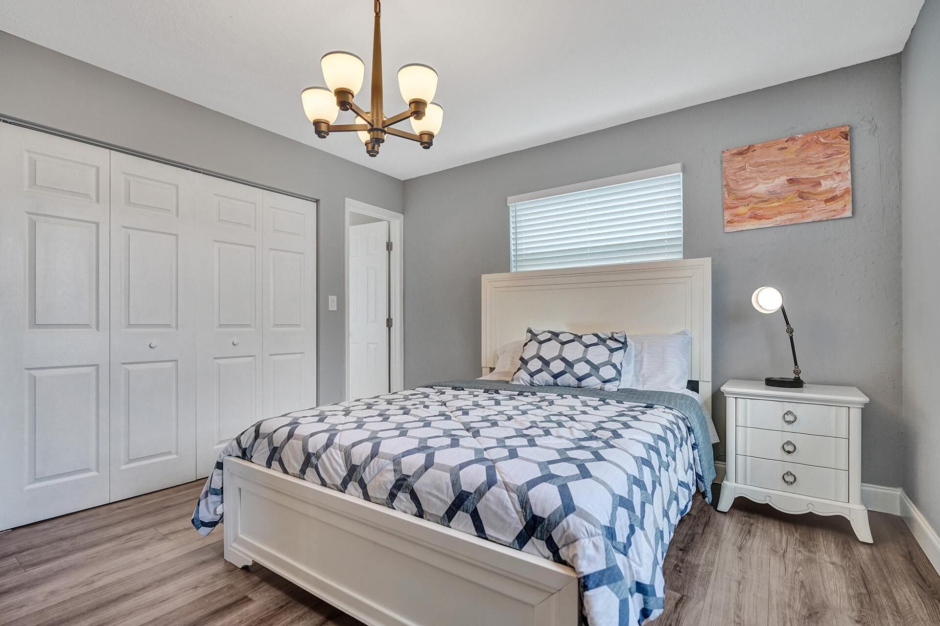 Une chambre à coucher comprenant un lit, une table de chevet, une lampe fixée au mur, une fenêtre avec des stores et une porte.