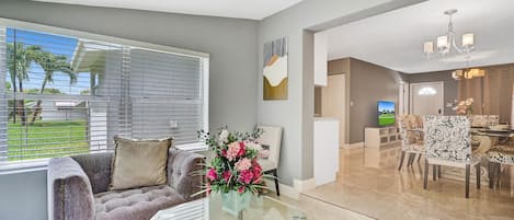 A living room with a grey sofa, a glass coffee table, and a vase of flowers. There is a dining area with patterned chairs and a chandelier.